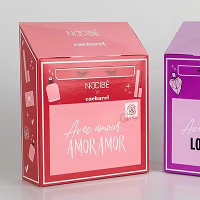 Deux coffrets cubiques côte à côte. L'un est violet et l'autre rouge. Sur une de ses faces, il est écrit "Avec Amour... AMOR AMOR" en lettres scriptes.