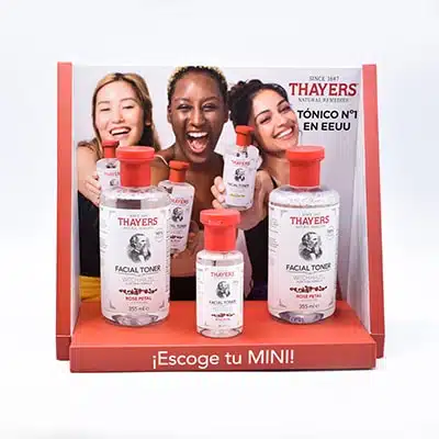Trois flacons contenant des produits de beauté sont disposés côté à côté dans un PLV en carton. Le carton possède plusieurs faces oranges. Sur l'une d'elle, on peut voir trois jeunes femmes souriantes.